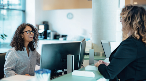 Two people talk across a desk with a computer in a bright office.