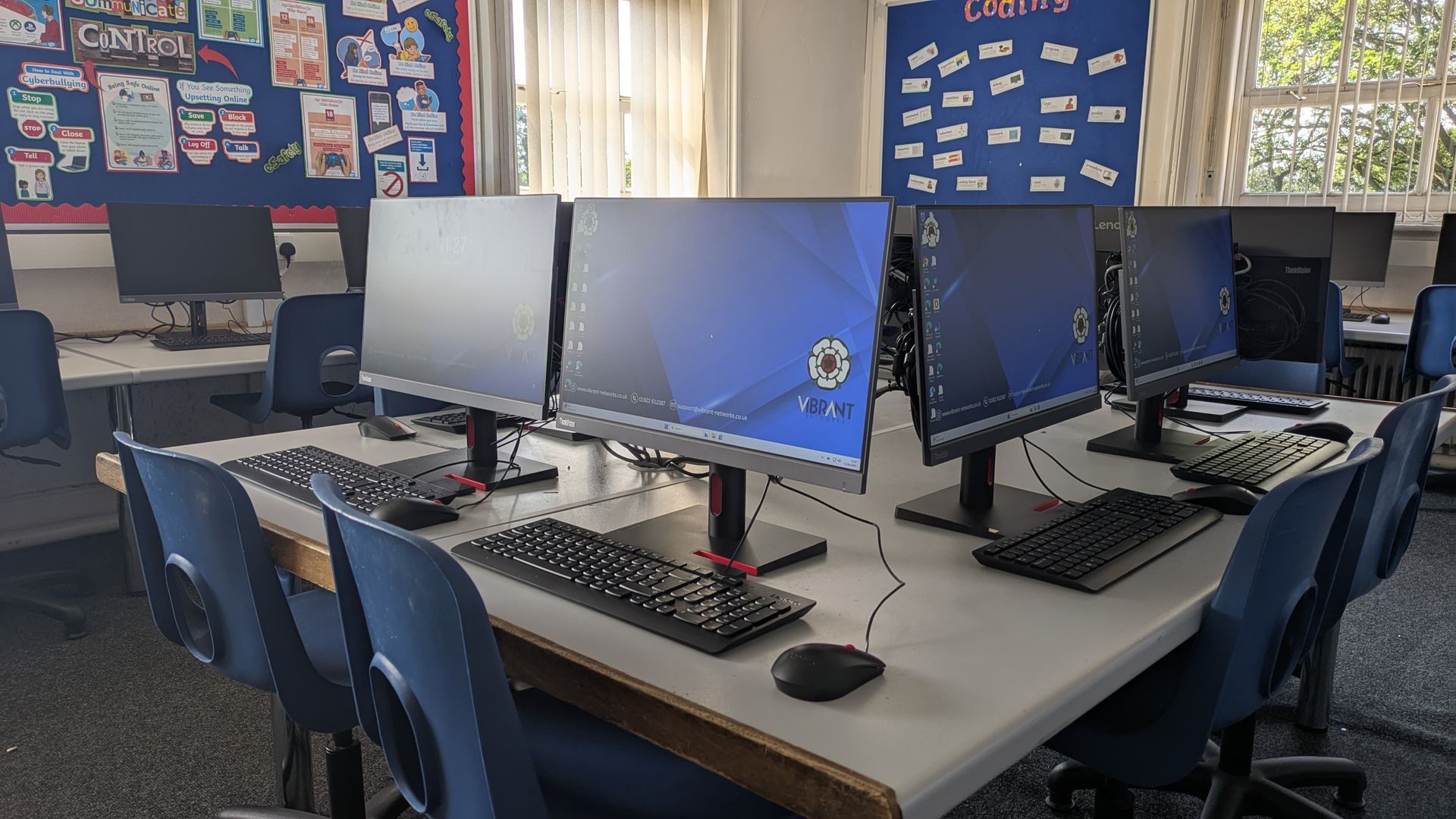 School computer lab with multiple desktop monitors, keyboards, and blue chairs arranged around tables.