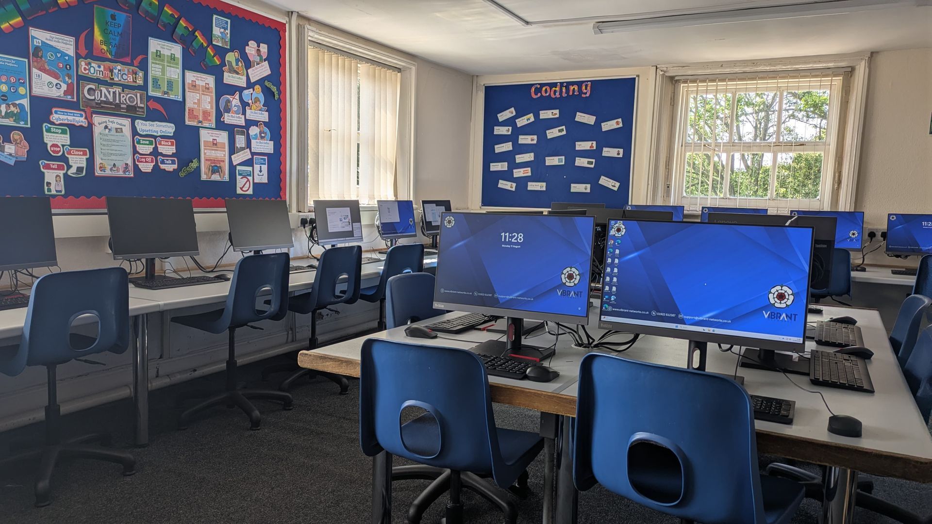 School computer lab with blue desks, desktop monitors, and wall displays in a bright classroom