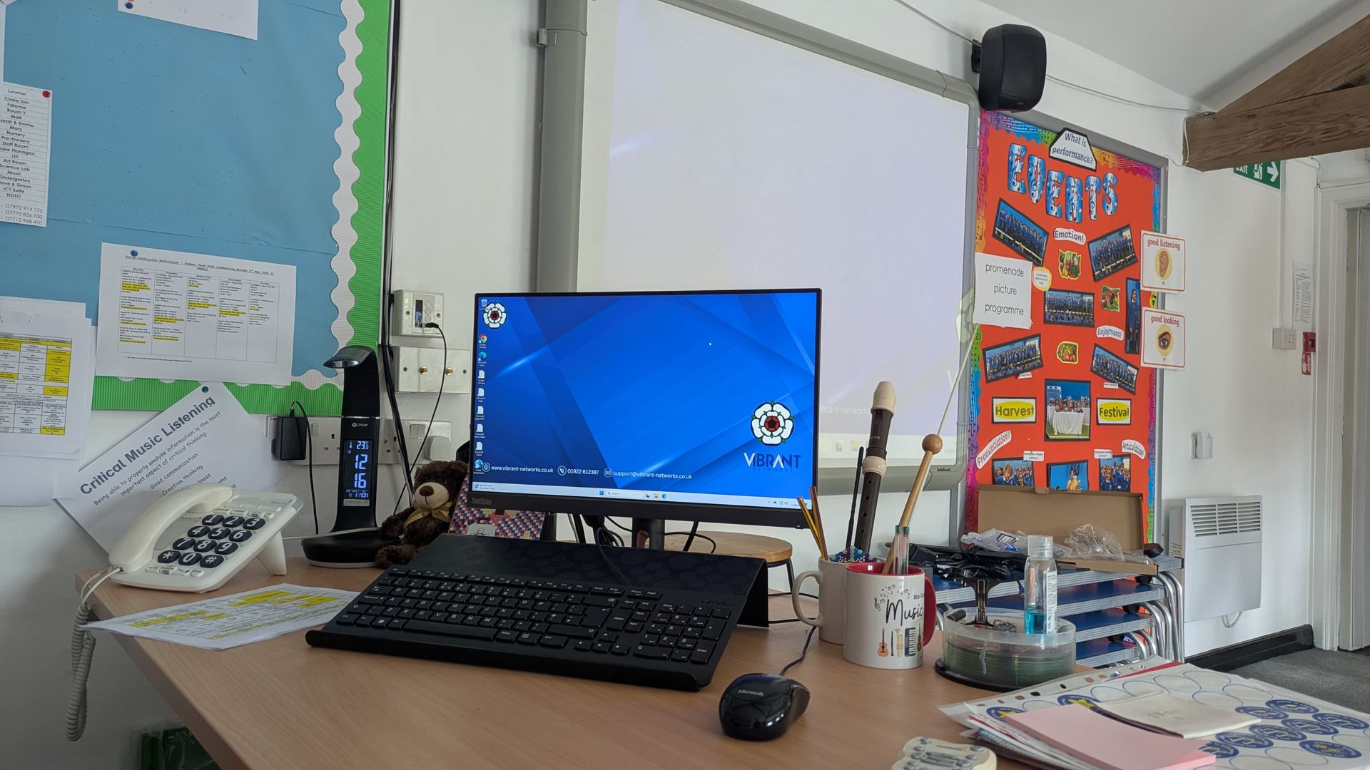 Desk with computer monitor, keyboard, mouse, and office supplies in a colourful classroom.