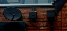Satellite dish and Zyxel Wi-Fi router mounted on a brick wall beneath a roofline.