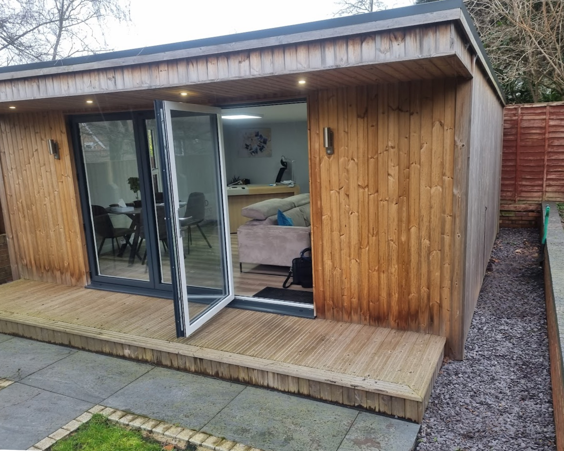 Wooden garden cabin with open glass doors, showing a furnished interior and a small deck outside.