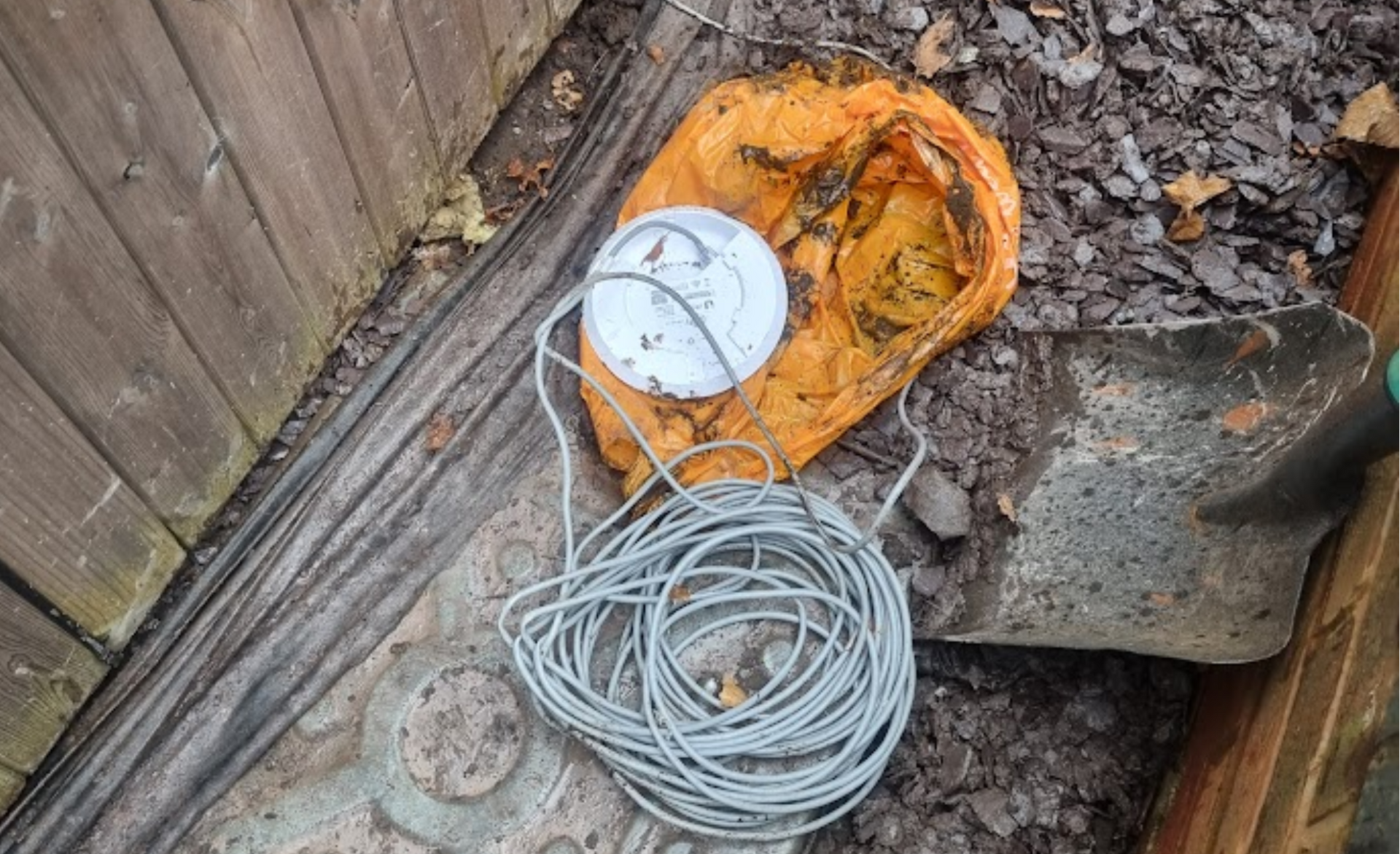 Coiled cable, white round Wi-Fi router, and orange extension cord on muddy ground beside a fence and shovel