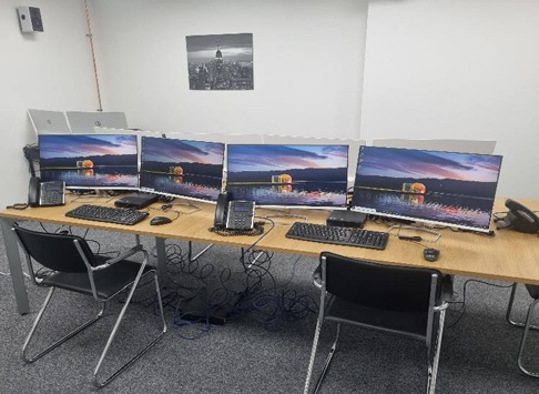 Row of four computer workstations on a wooden desk in a plain office room
