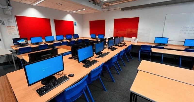 School computer lab with rows of desktop monitors on wooden desks and bright red wall panels.