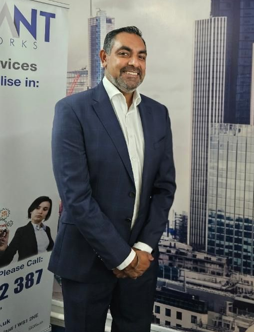 Man in a navy suit standing in front of a cityscape banner, smiling with hands clasped.