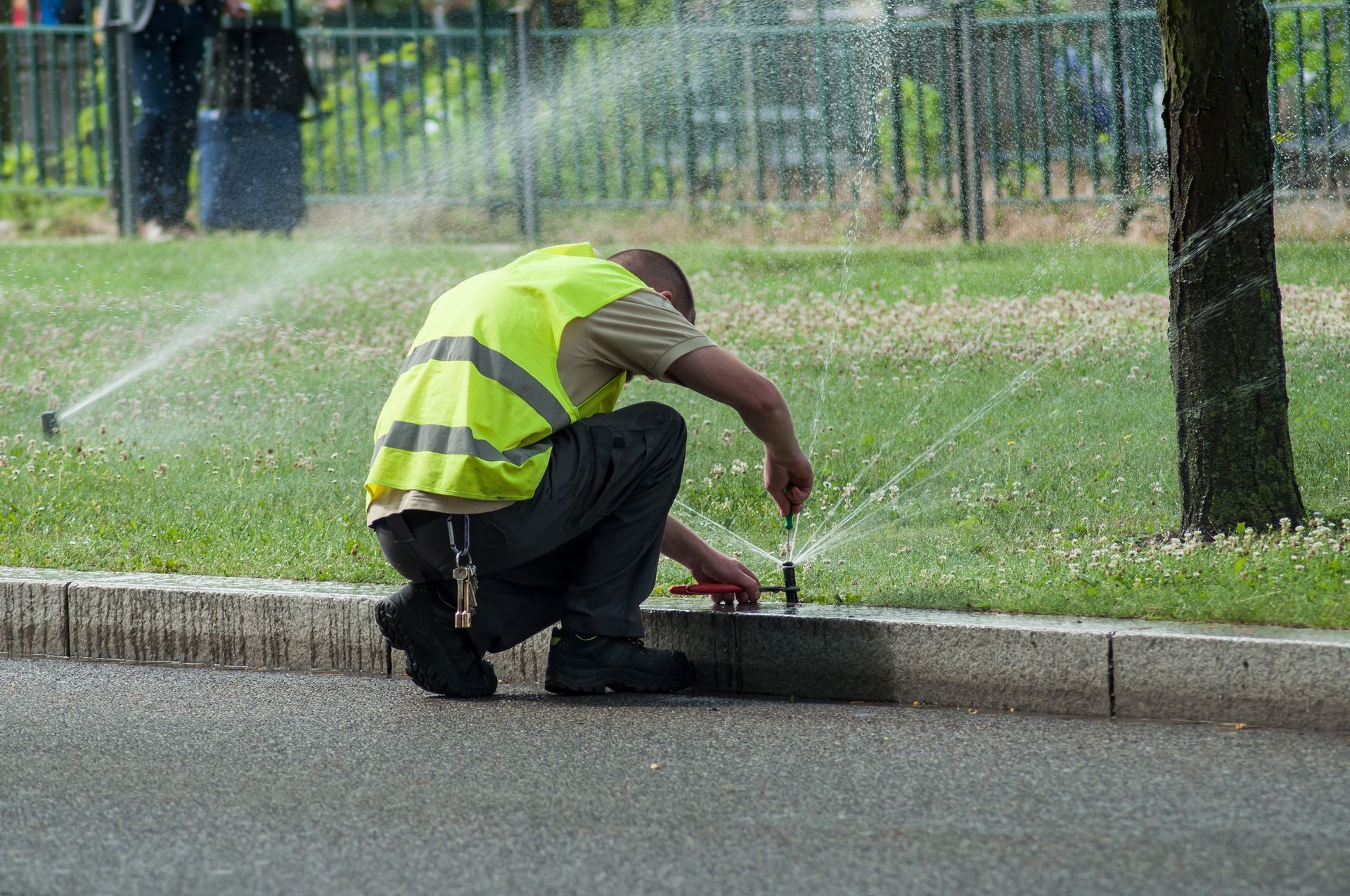 Worker in a neon safety vest kneels by a curb, adjusting a sprinkler on a wet roadside.