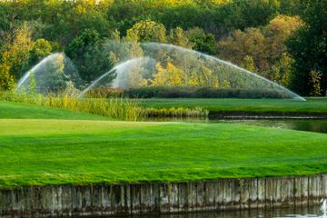Green lawn with water sprinklers spraying arcs near a pond and trees in the background