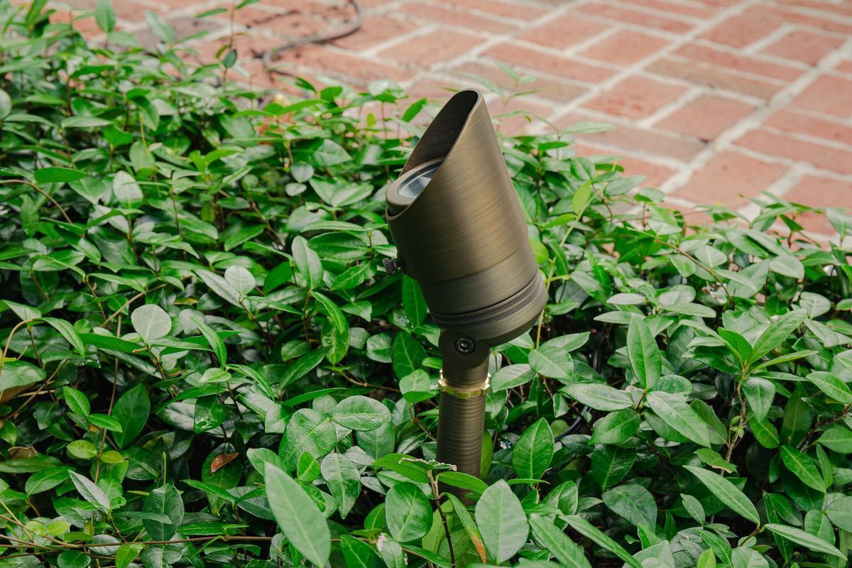Metal garden spotlight among green shrubs beside a brick patio.