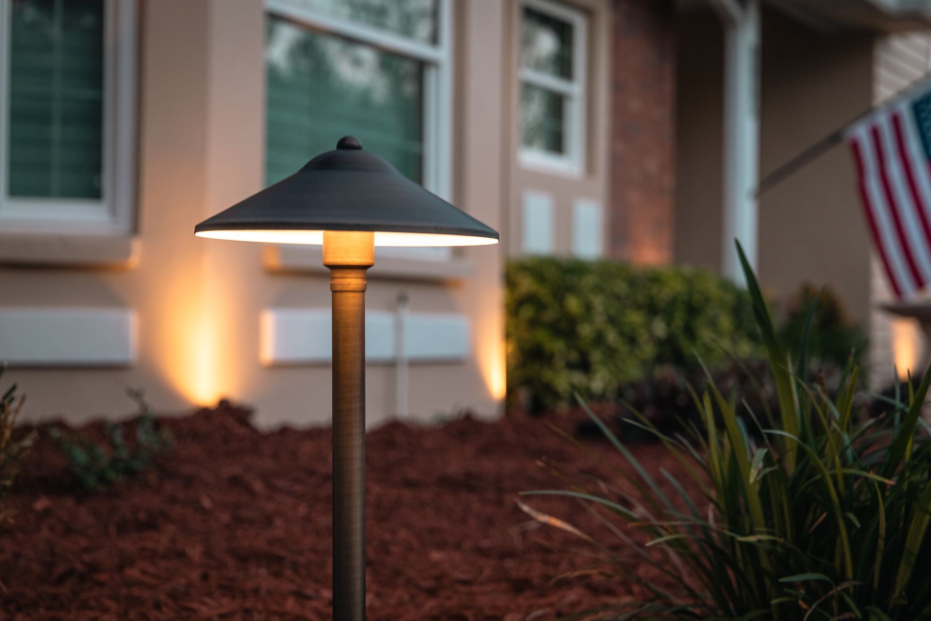 Lit garden pathway lamp glowing beside a brick house and shrubs at dusk