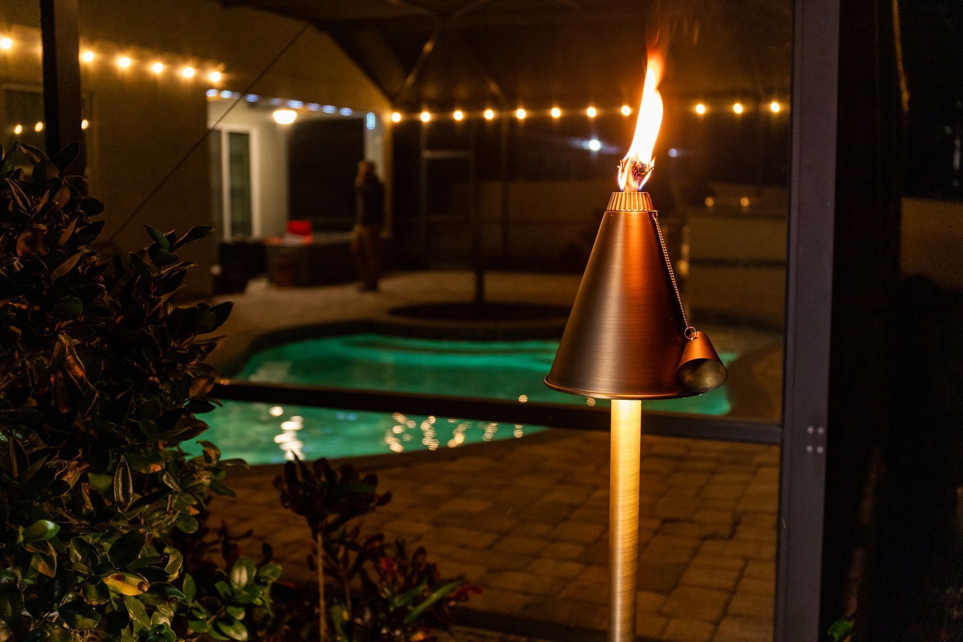 Lit patio with tiki torch beside a lit swimming pool at night