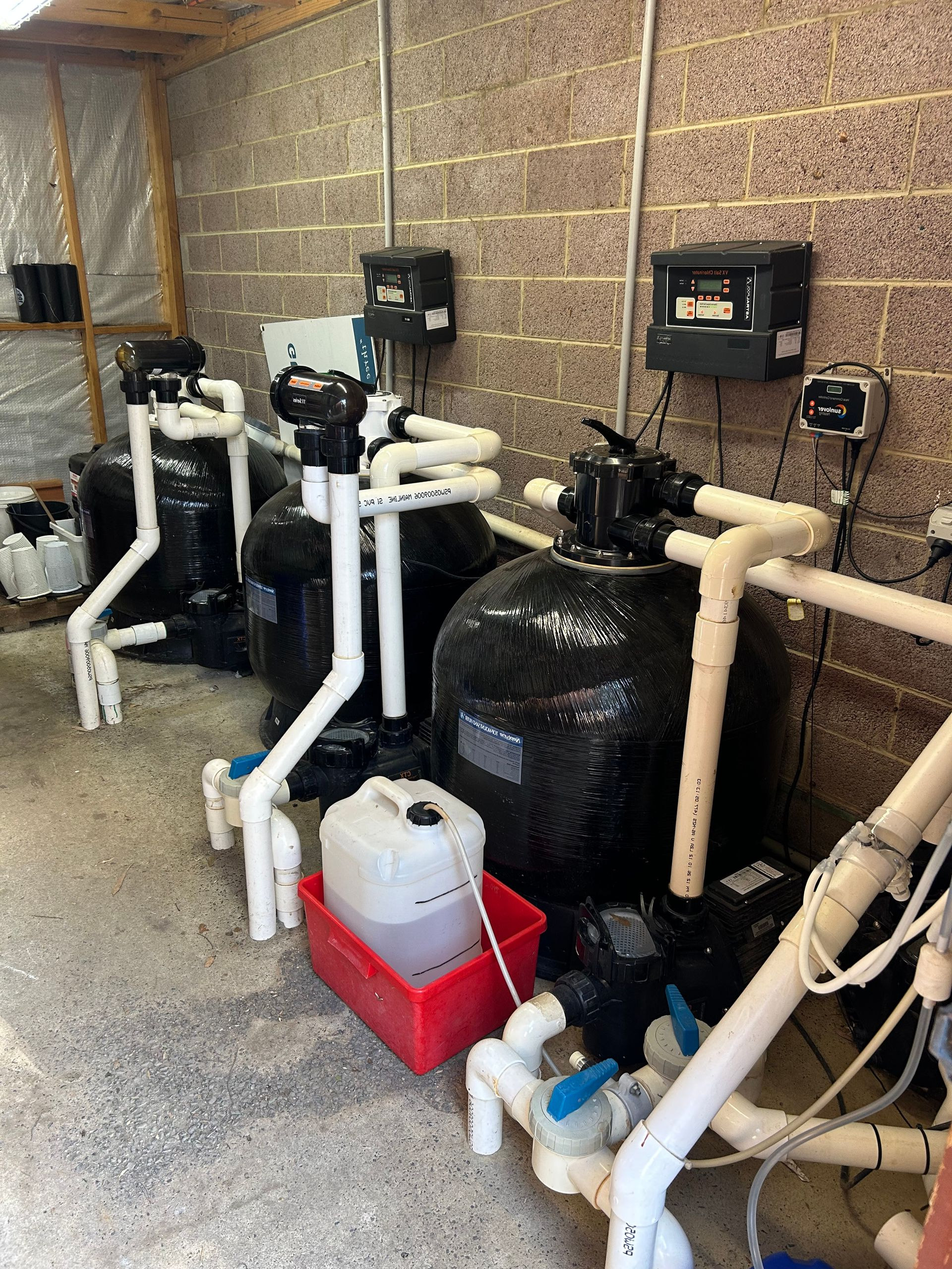 Row of black pool pumps and white PVC piping beside a brick wall in a utility room