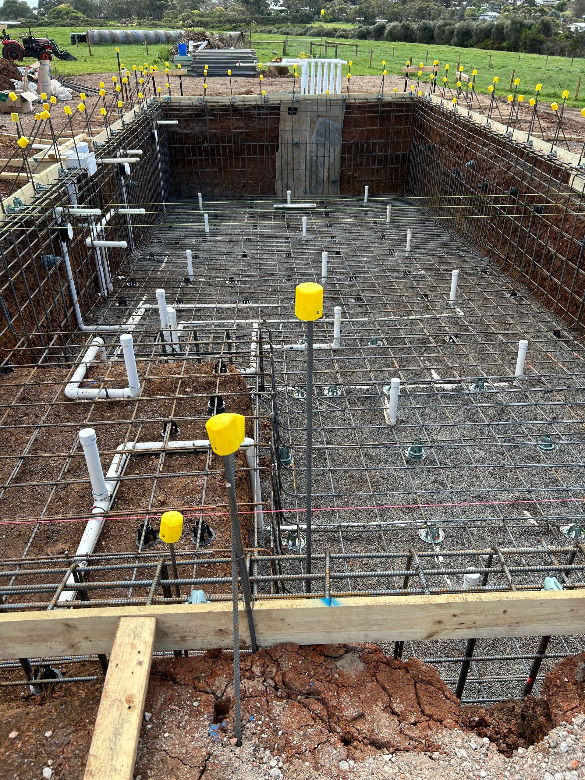 Large rectangular concrete foundation excavation with rebar grid and yellow pipe caps at a construction site