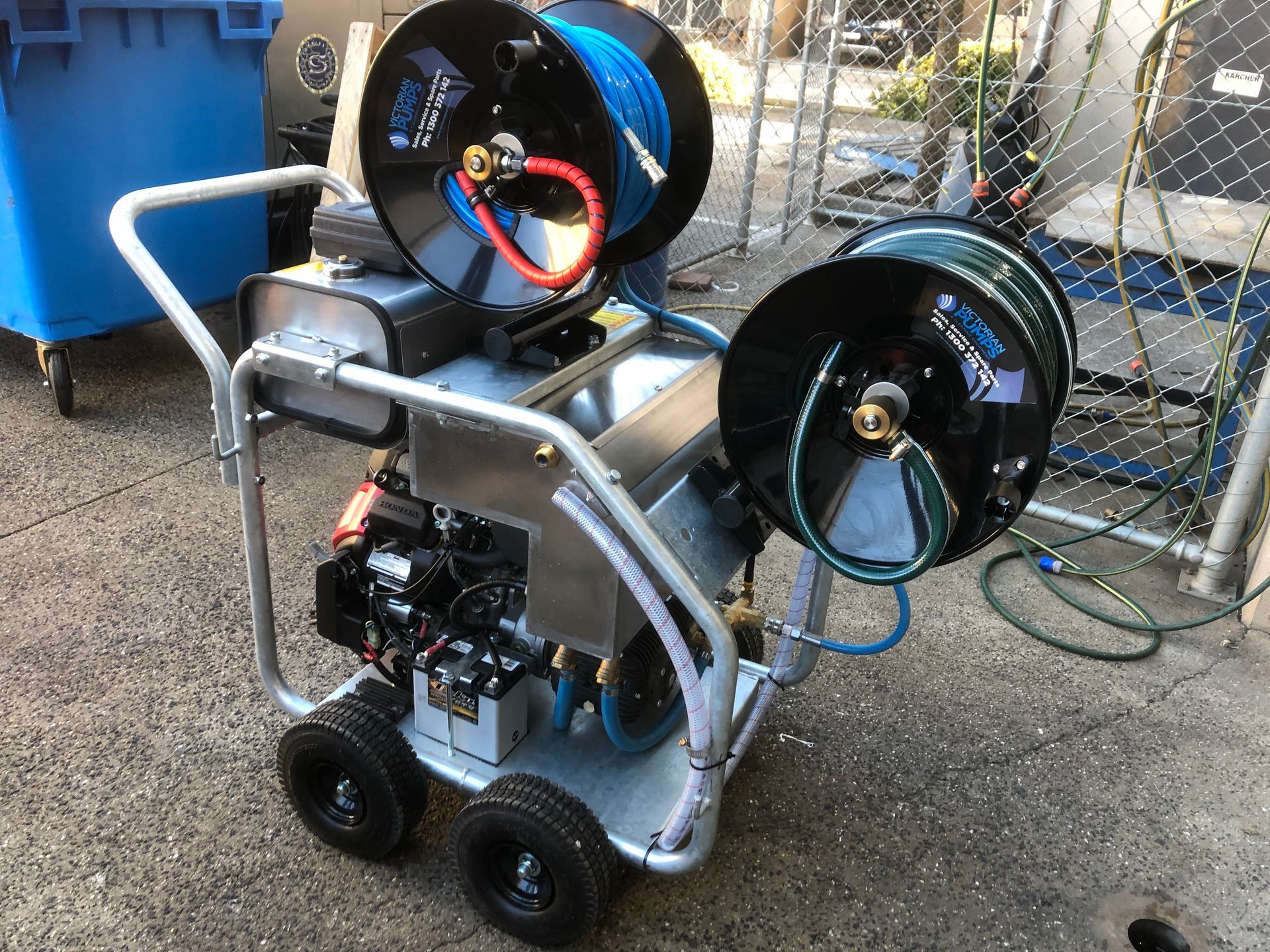 Portable machine on a wheeled frame with two large black hoses or reels, outdoors on pavement.
