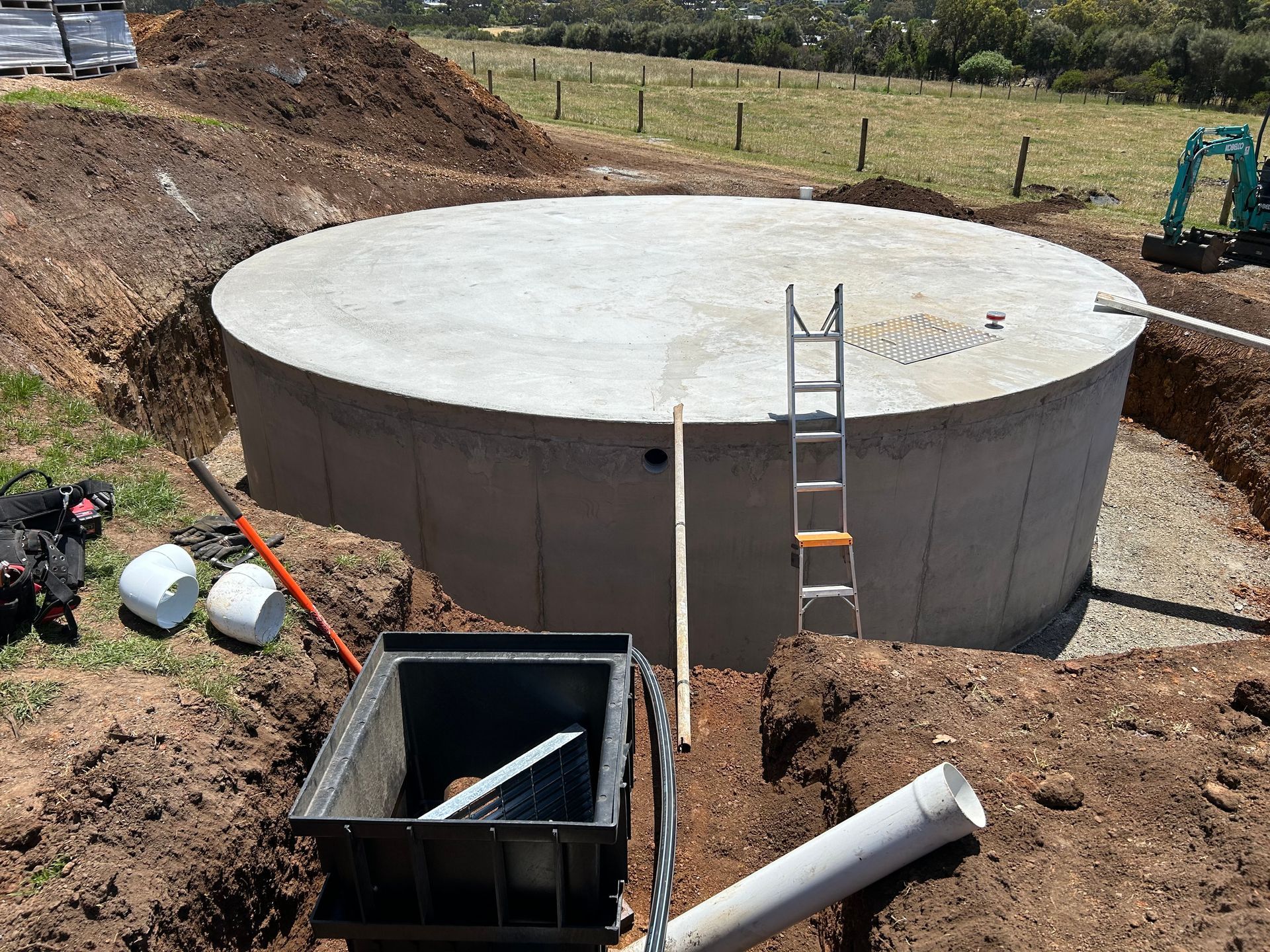 Concrete circular tank at a construction site with a ladder, excavator, and dirt trench nearby
