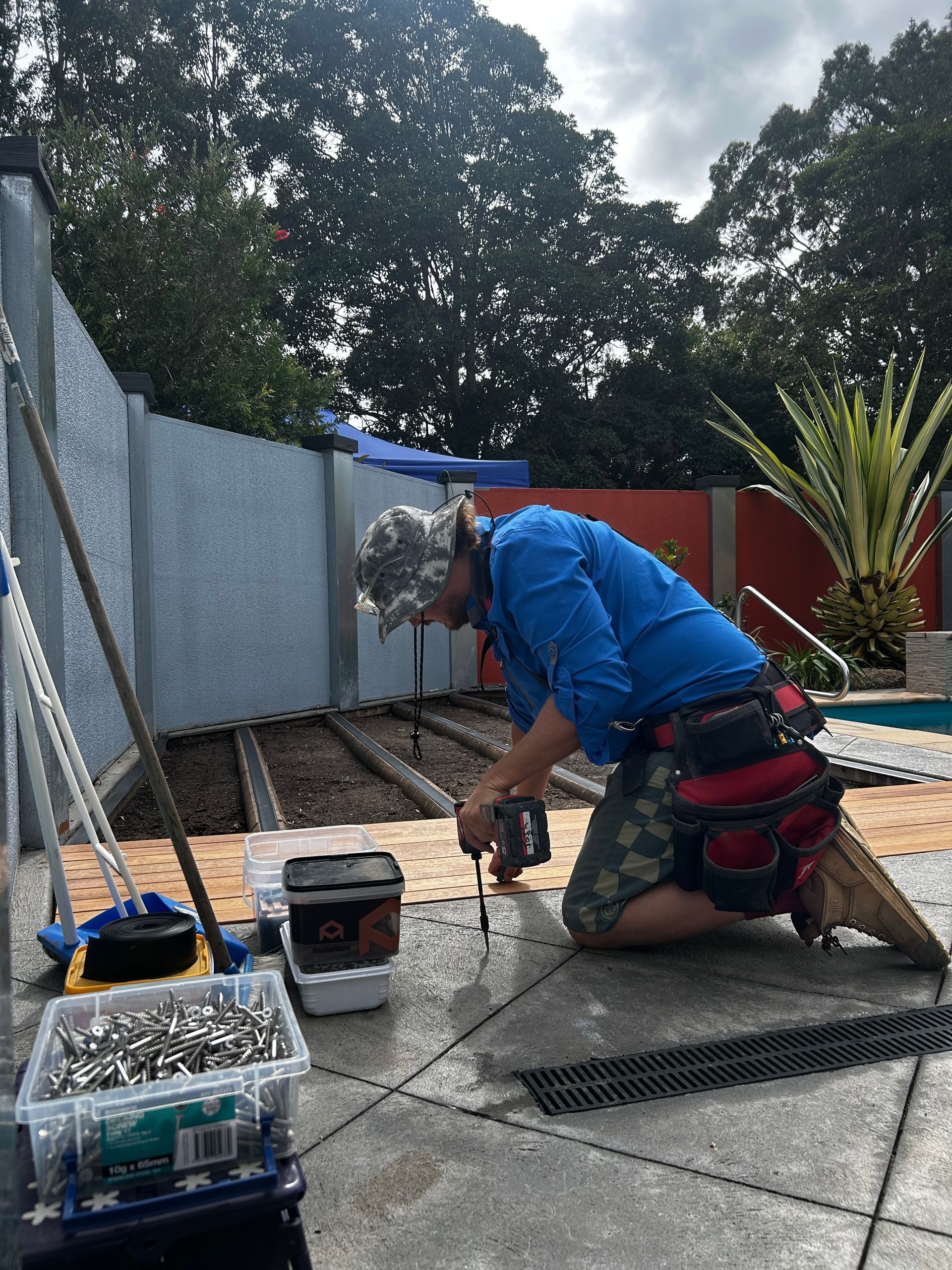 Worker in blue shirt kneeling on a patio, using a tool beside buckets and supplies near a red fence.