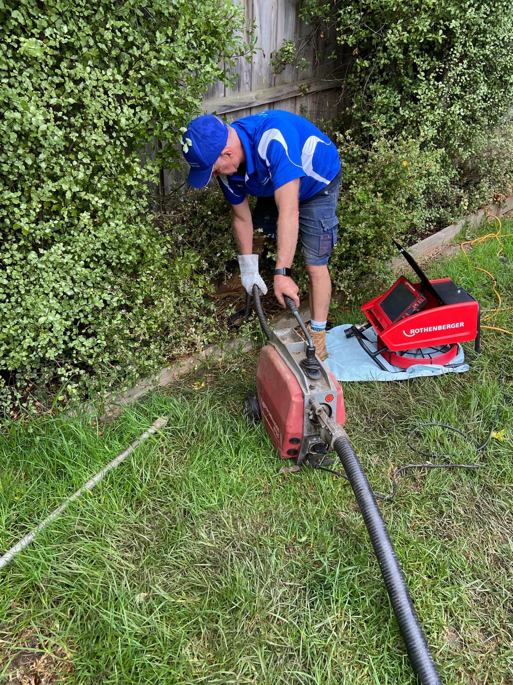Worker in blue uniform using a gas-powered trimmer along a grassy hedge, with a red tool case nearby.