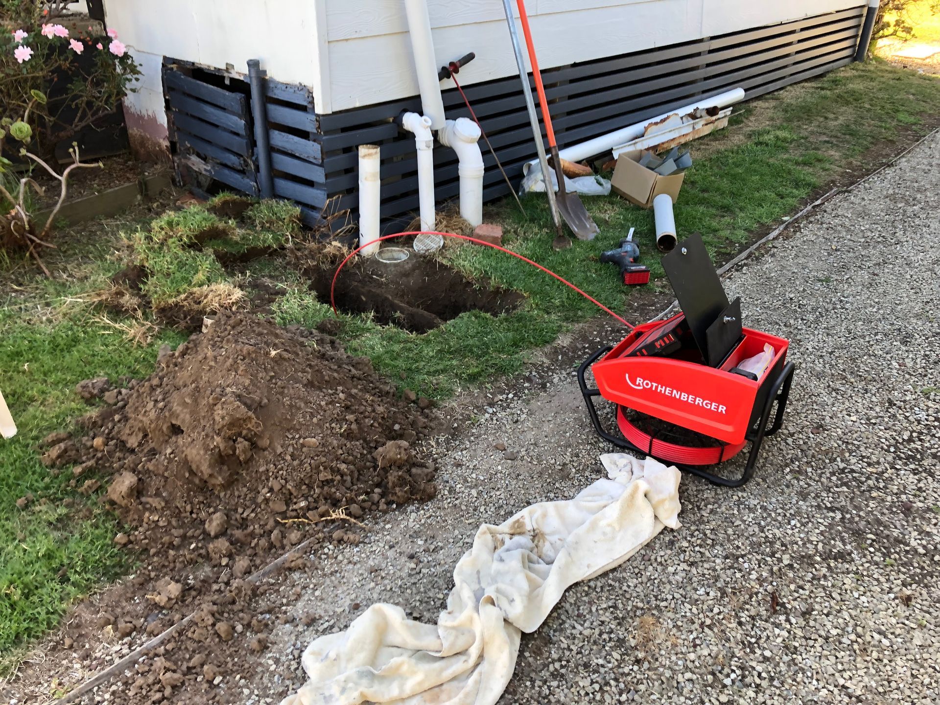 Outdoor yard excavation with dirt pile, white pipes, red blower, and gravel beside a house.