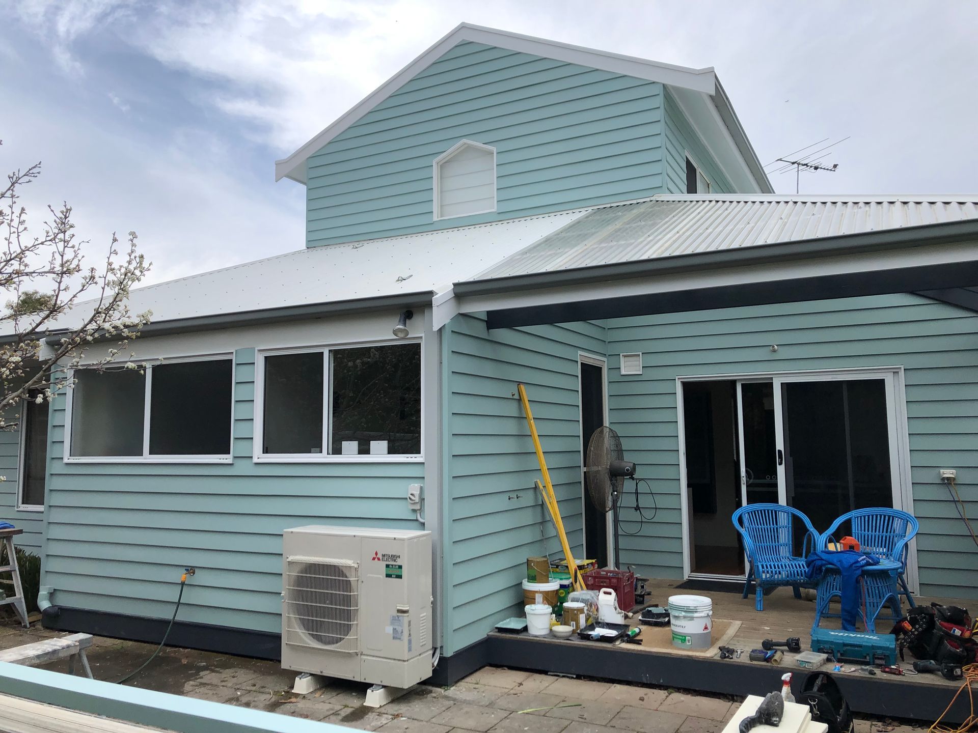Light blue house exterior with a small porch, patio chairs, and gardening tools on a cloudy day