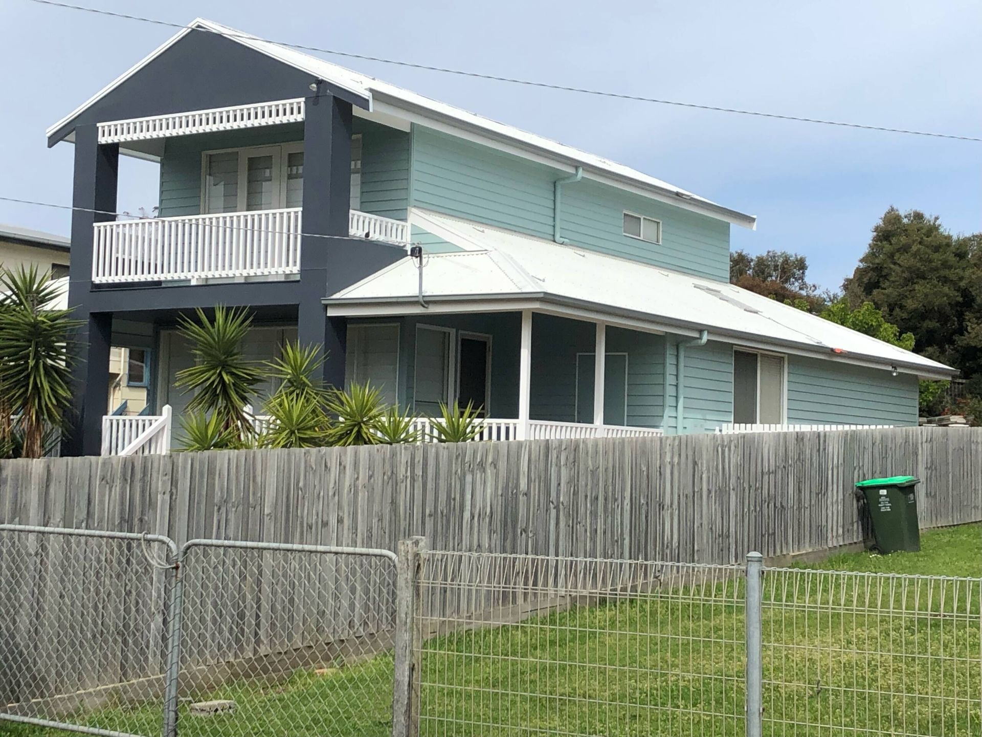 Two-story pale blue house with white trim and a fenced yard on a sunny day