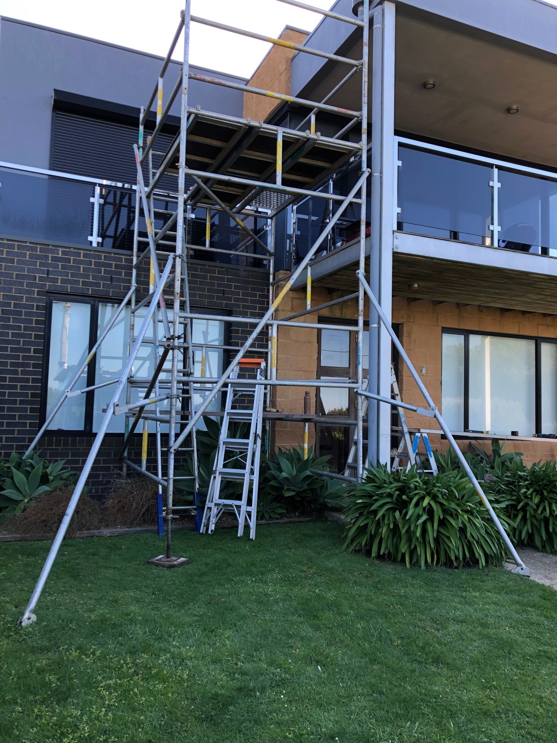 Scaffolding and ladders outside a brick apartment building with a balcony and lawn