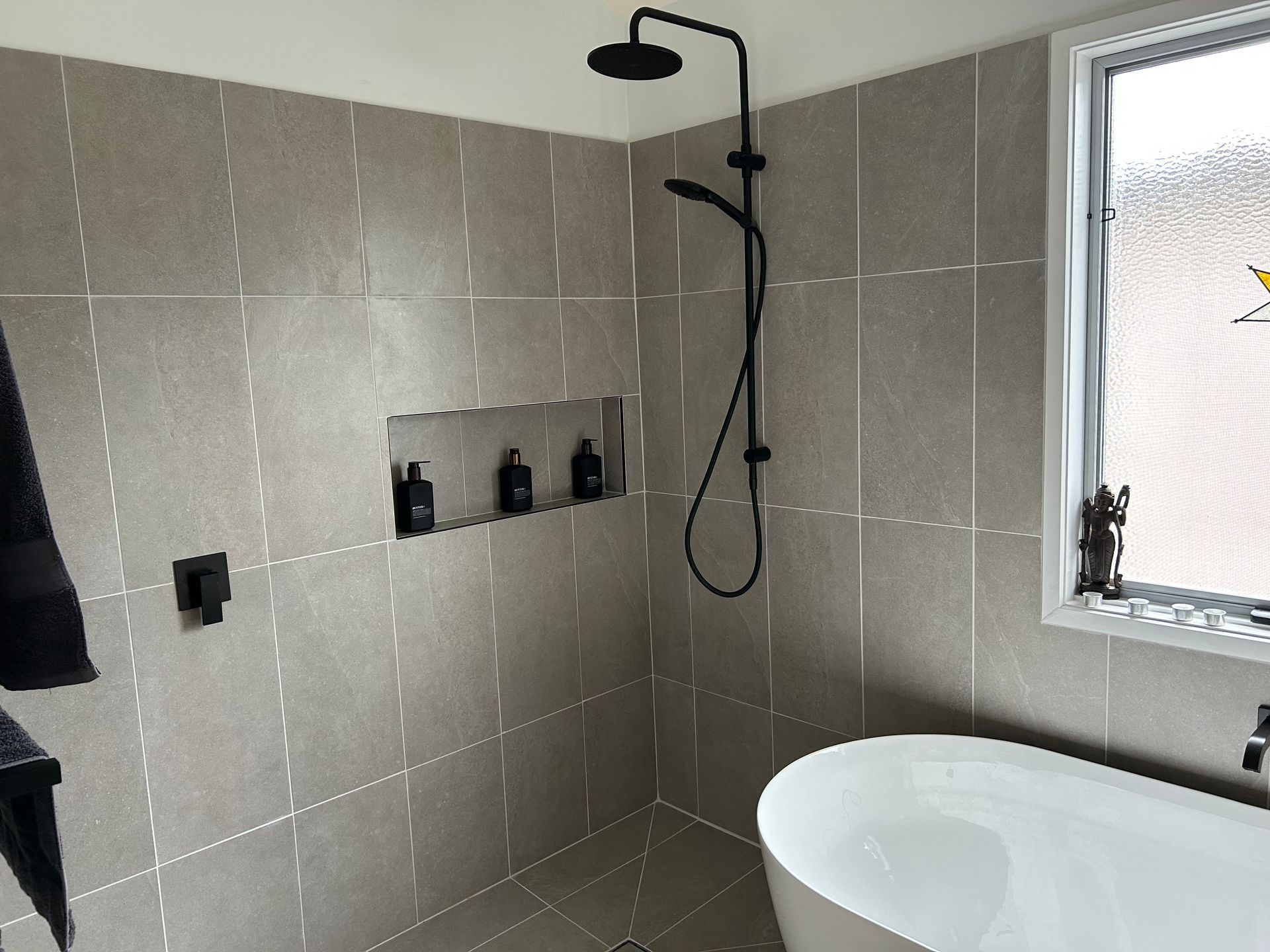 Modern bathroom shower area with gray tiled walls, black shower fixtures, and a white sink by a frosted window