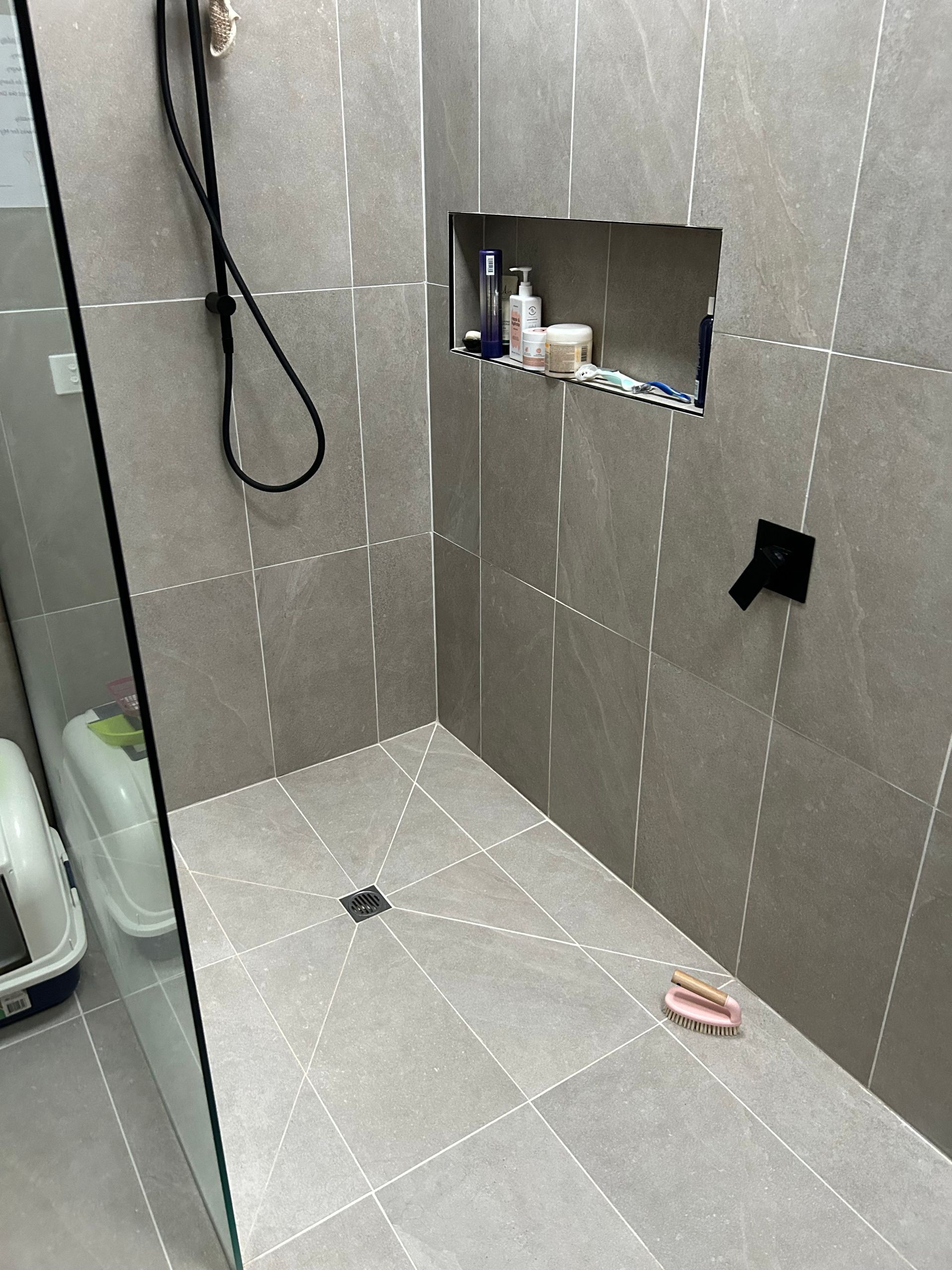 Gray tiled shower stall with recessed shelf, hanging hose, and toiletries on the floor.