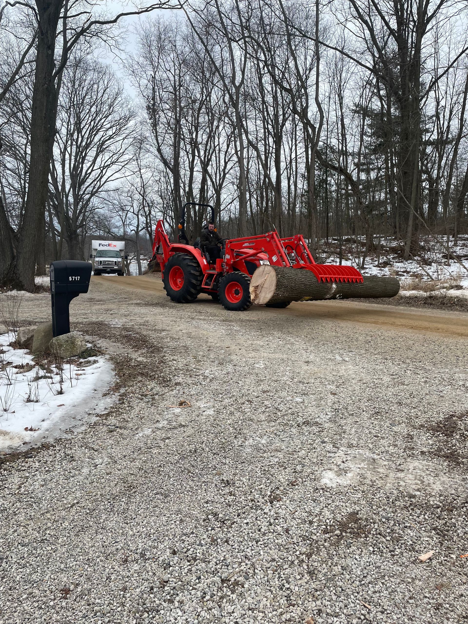 A red tractor is carrying a large log in a gravel driveway.
