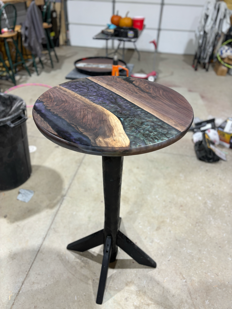 A round table with a live edge wooden top and custom epoxy inlay on a black base is sitting on a concrete floor.
