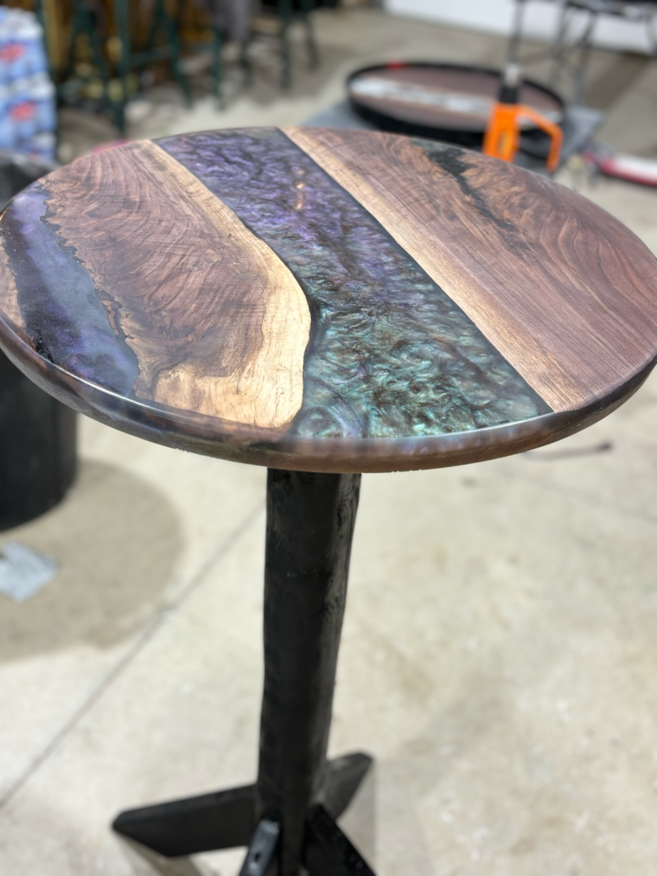 A round table with a live edge wooden top and custom epoxy inlay on a black base is sitting on a concrete floor.
