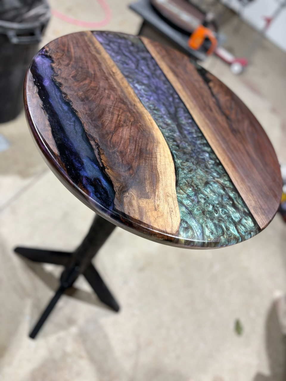 A round table with a live edge wooden top and custom epoxy inlay on a black base is sitting on a concrete floor.
