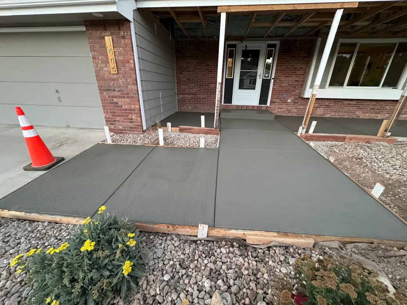 Newly poured concrete walkway leading to a house entrance. A traffic cone and gravel are also visible.