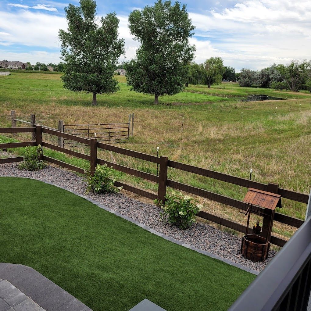 Green lawn next to a brown wooden fence, and field, with trees, blue sky.