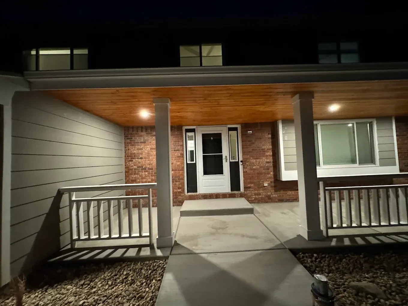 Front porch of a home at night. Lit doorway centered between support columns, brick wall, and wood ceiling.