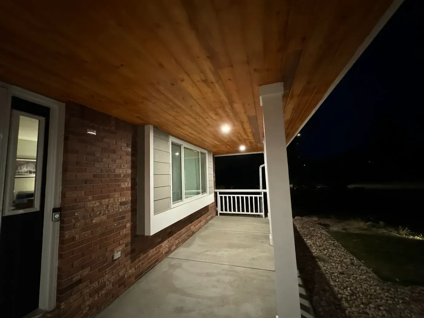 Covered porch with wood ceiling, brick wall, white trim, and a small railing.