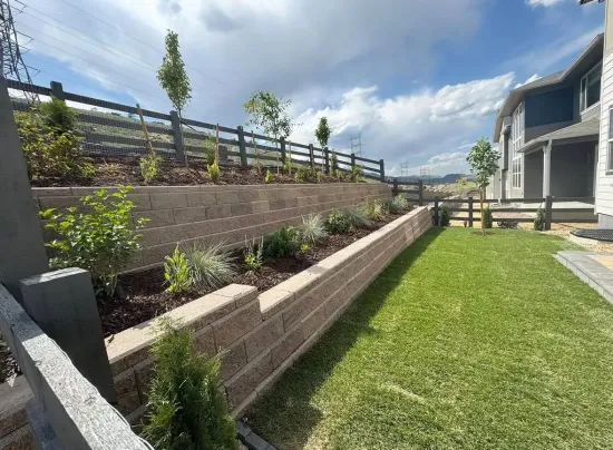 A backyard with tiered retaining walls, green grass, and a wooden fence under a cloudy sky.