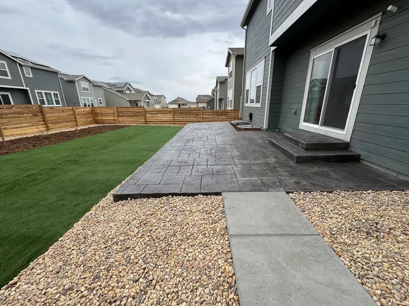 Backyard with gray patio, artificial grass, gravel, and wooden fence. House with sliding glass door.