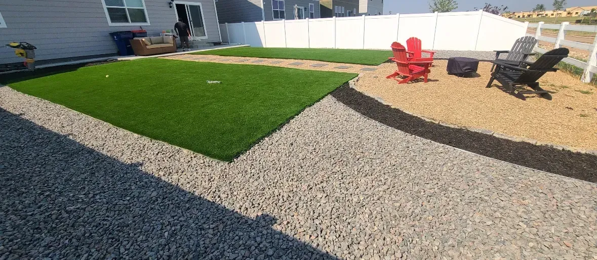 Backyard with artificial grass, gravel pathway, fire pit, and chairs.