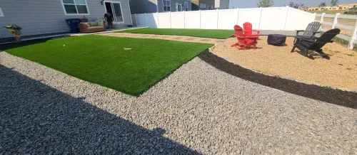 Backyard with green artificial turf, gravel paths, and a seating area with red and black chairs.