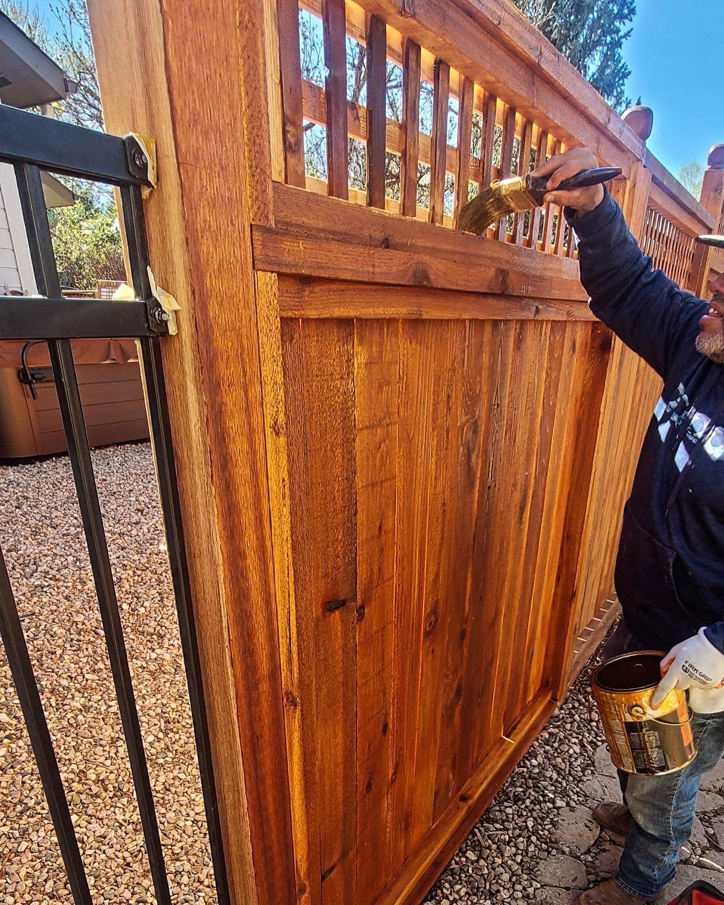 Person staining a wooden fence with a brush, holding a can of stain outdoors.