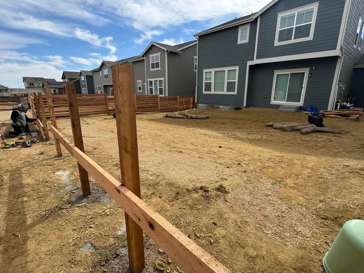 A partially built wooden fence in front of several two-story houses on a dry, dirt yard under a cloudy sky.