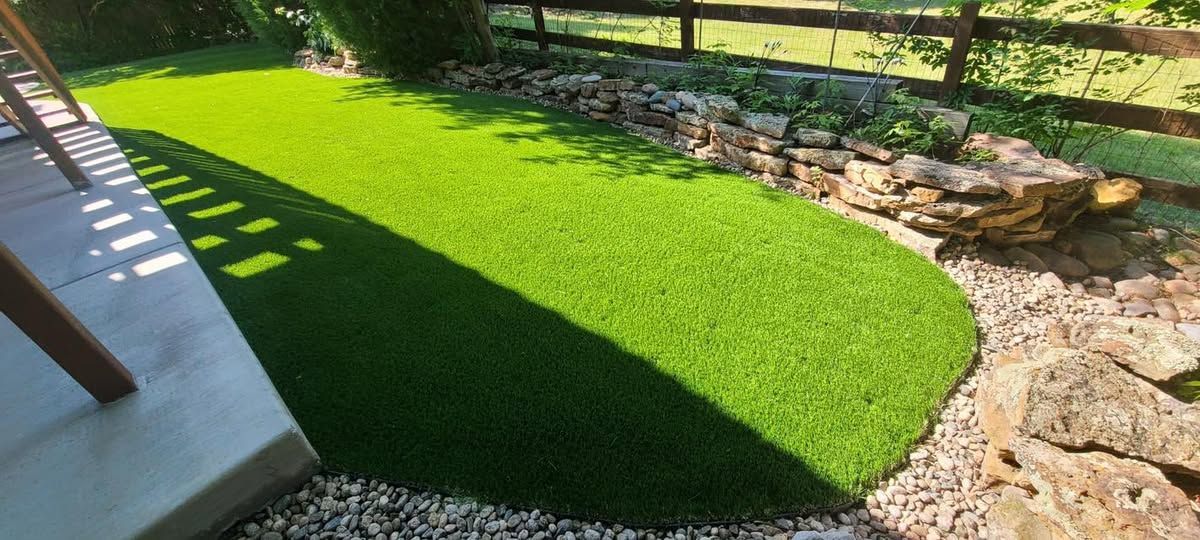 Lush green artificial turf surrounded by stones, a wooden fence, and a patio area.