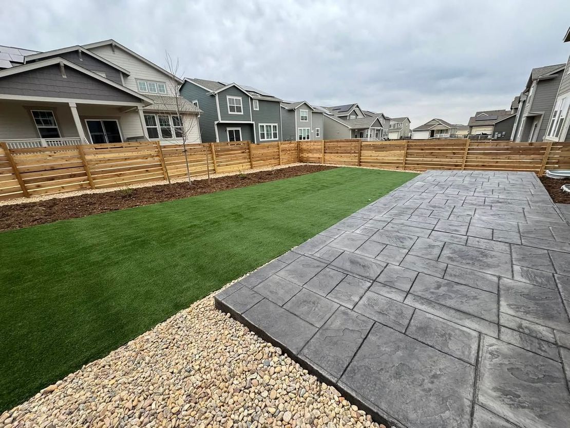 Backyard with stamped concrete patio, green turf, gravel, and wooden fence. Row of houses in the background.