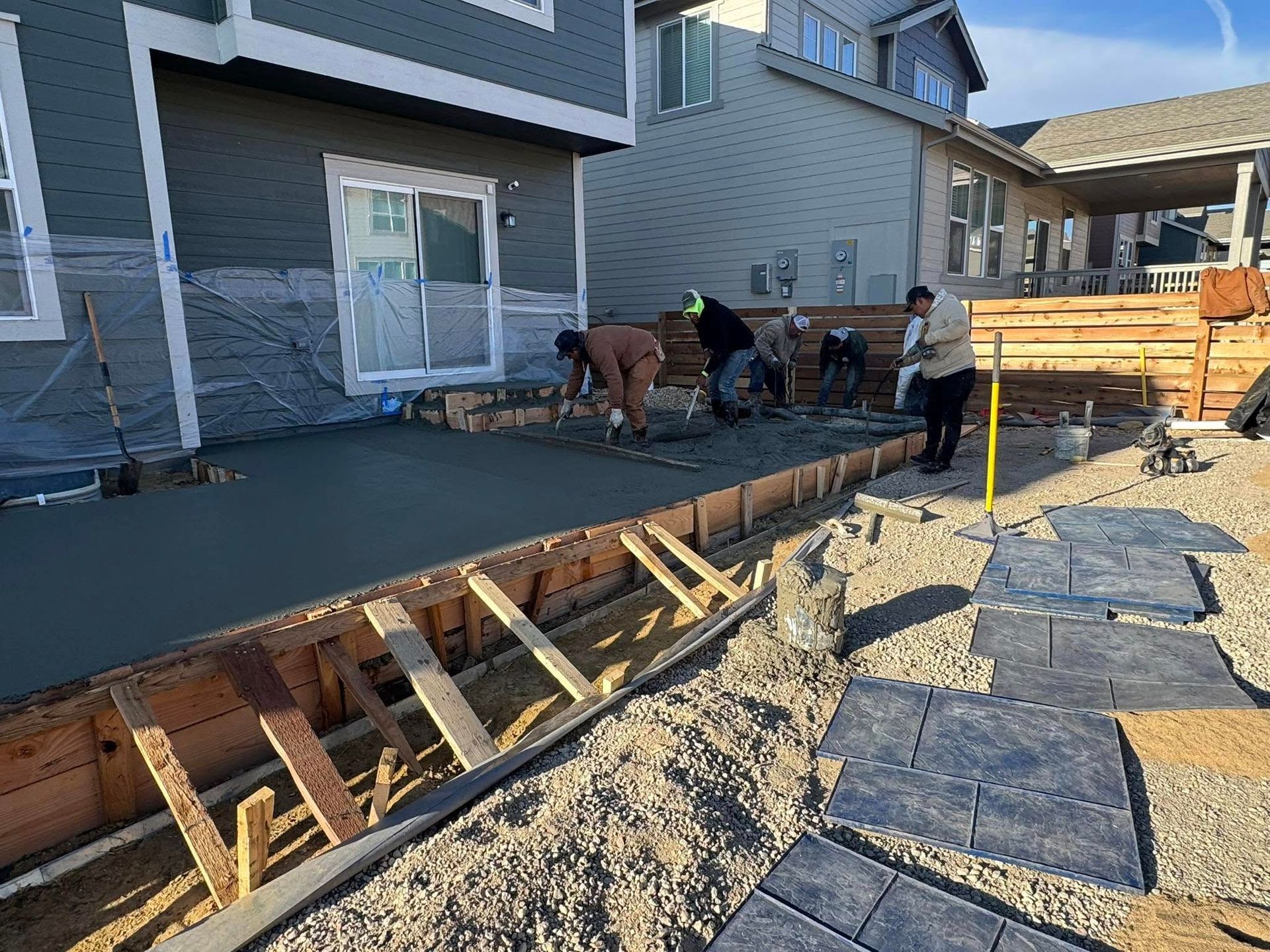 Construction workers leveling wet concrete for a patio, next to a house with a sliding door.