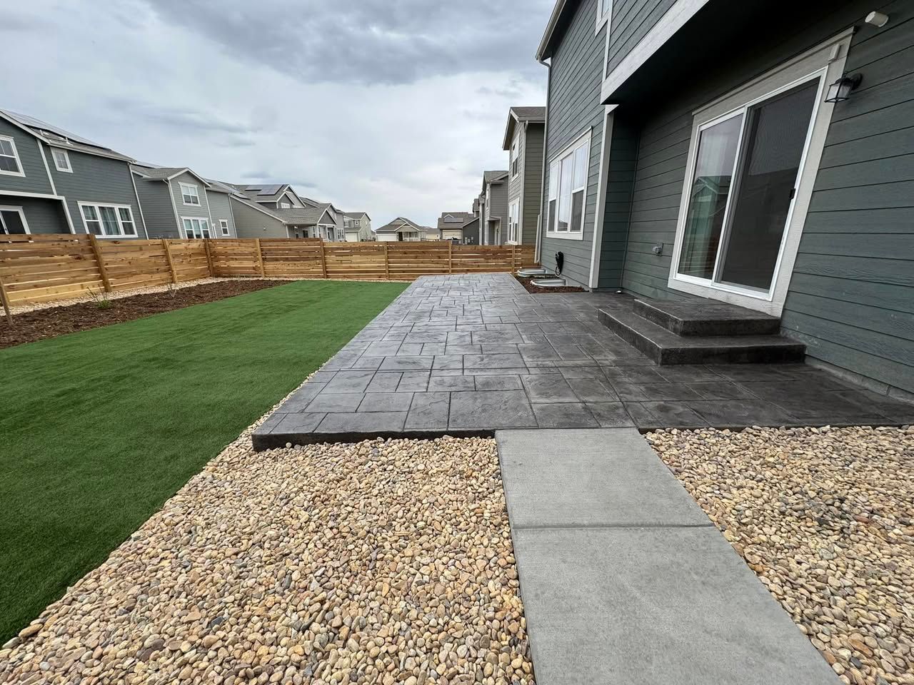 Backyard patio with stamped concrete, a gravel border, and a green lawn, adjacent to a house with a sliding glass door.