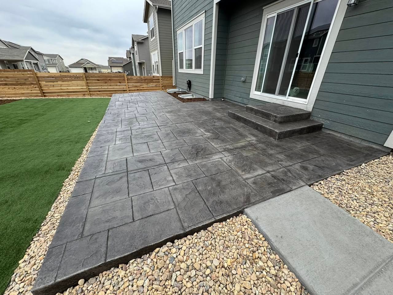 Gray stamped concrete patio with steps leading to a sliding glass door, edged with gravel and a lawn.