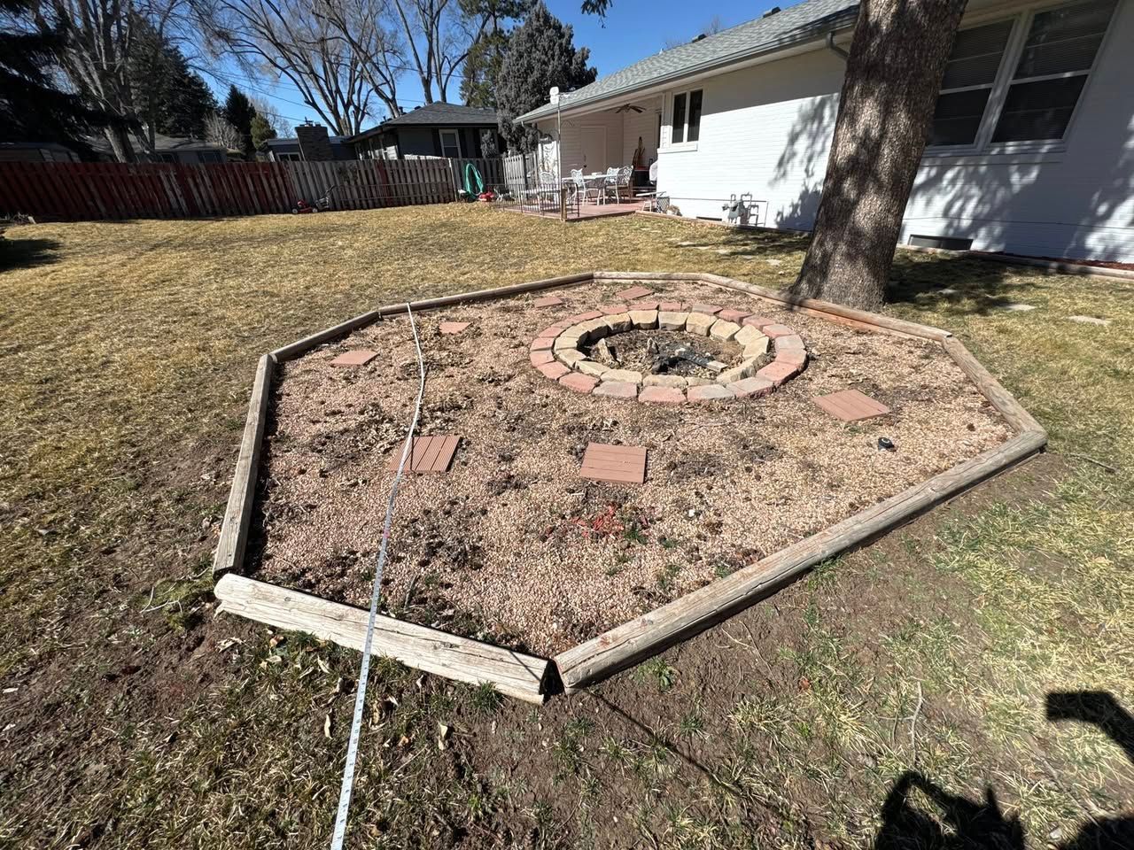 Backyard fire pit surrounded by tan gravel and brick with a house and tree in the background.