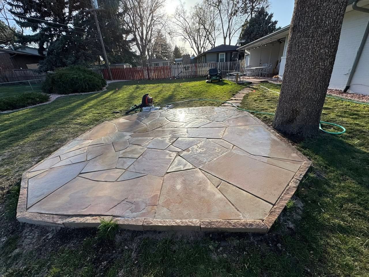 A stone patio in a backyard with a tree and green grass.