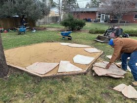 Man laying flagstone pavers in a sandy octagonal area in a backyard. Blue wheelbarrows and small excavator visible.