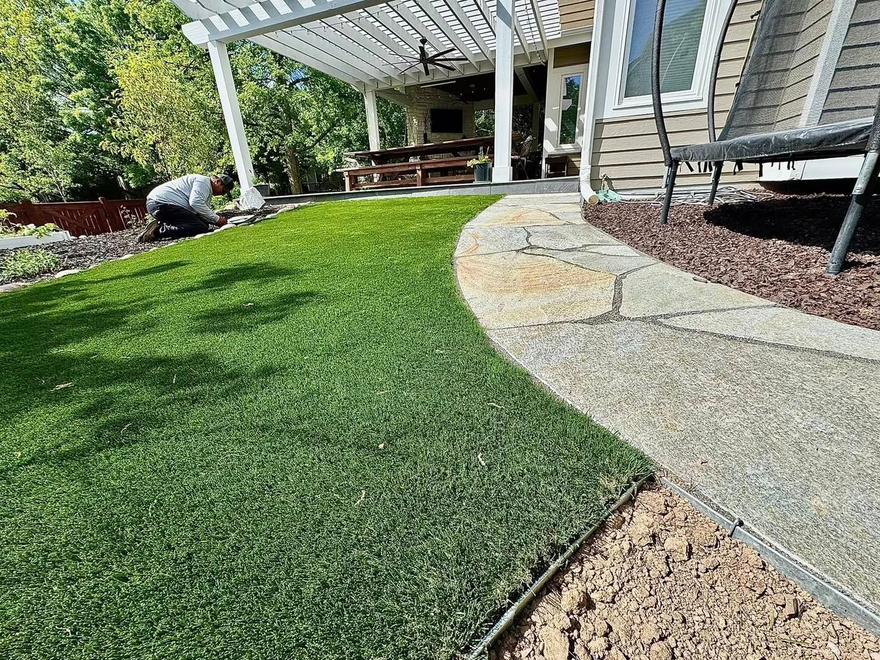 Green turf next to a stone path and mulch. A person works on the landscaping near a house with a pergola.